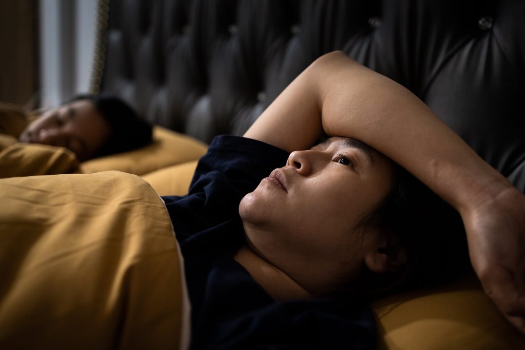 Asian woman lying in bed staring at the ceiling unable to fall asleep representing someone who would benefit from Insomnia Treatment in Arizona.