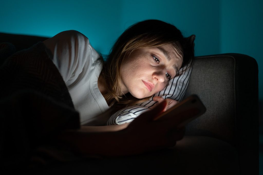 Young woman staring at her phone having difficulty falling asleep representing someone who would benefit from Insomnia Treatment in Arizona.
